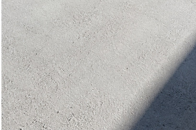 Macro photograph of the mineral-finish surface of PIEZA 001, showing its aggregate texture beneath a hard diagonal shadow.