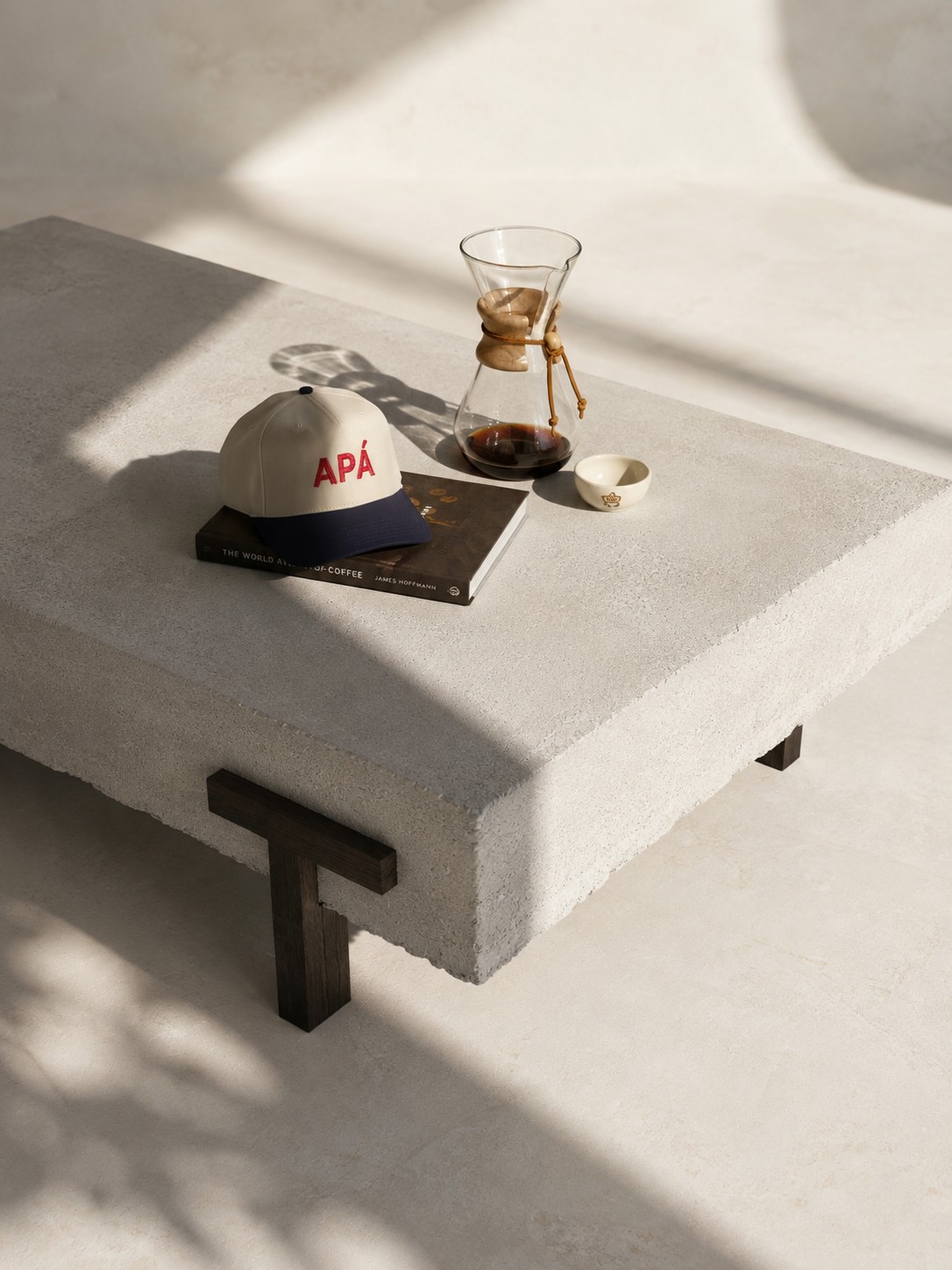 PIEZA 001 styled in use, with an APÁ cap resting on a coffee book, a Chemex of brewed coffee, and a small ceramic cup on the table top in raking sunlight.