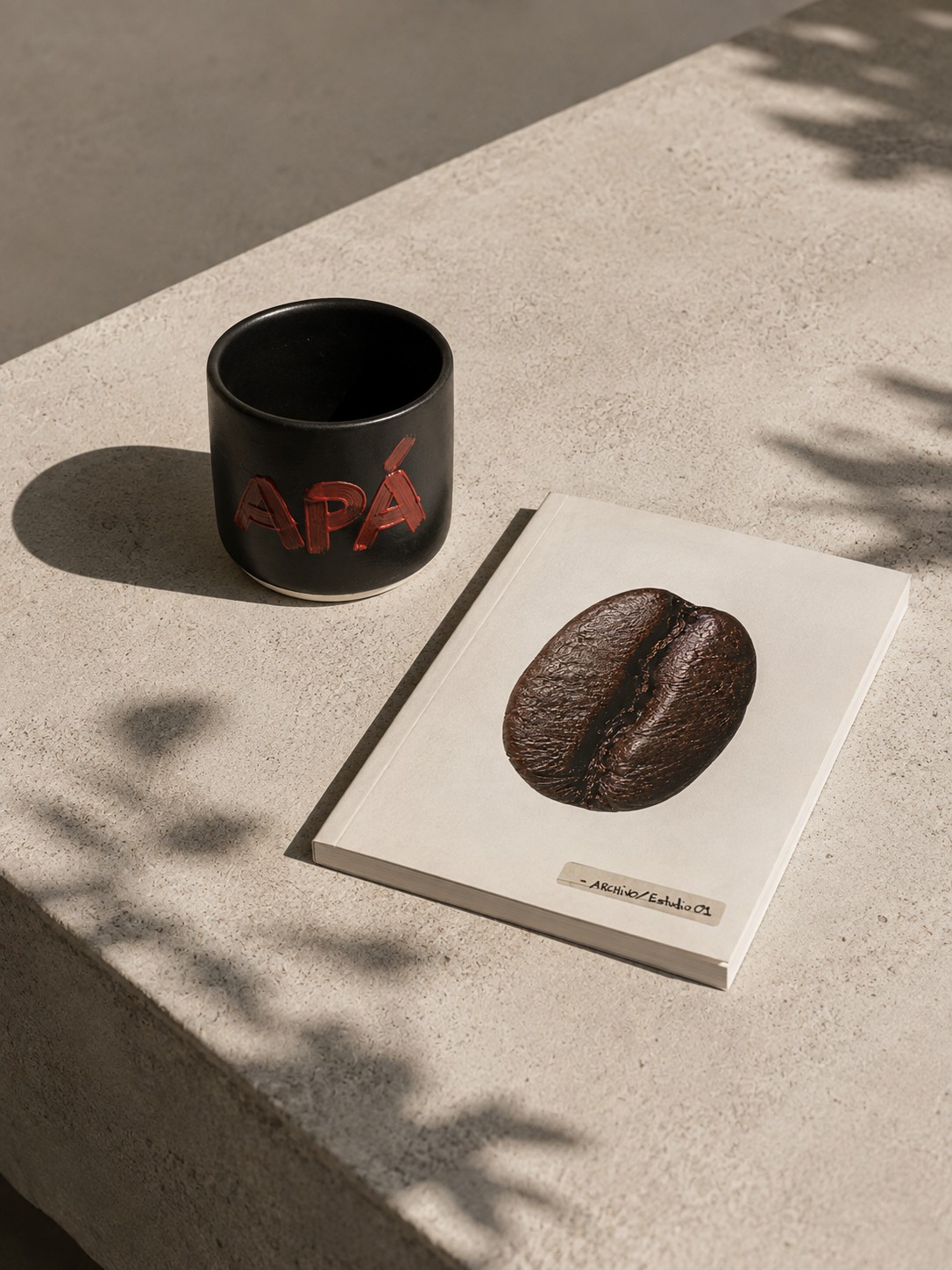 A black ceramic APÁ cup with red lettering beside the Pacamara ARCHIVO / ESTUDIO 01 lookbook, resting on the corner of the mineral-finish table in dappled light.